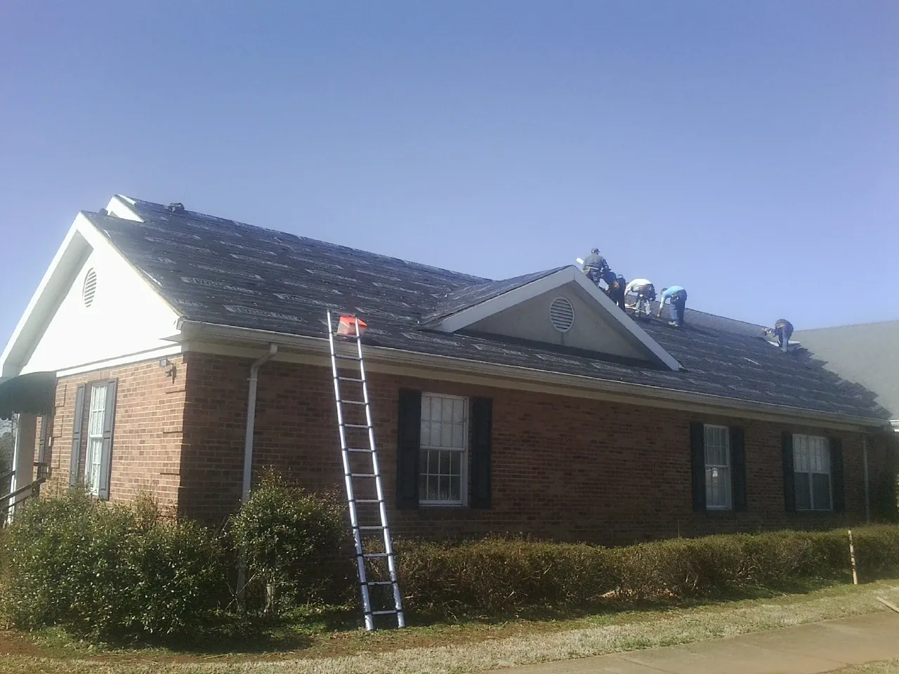 Crew tearing shingles off a brick ranch roof with a ladder set — Emergency Roof Repair work by our crew in Eva