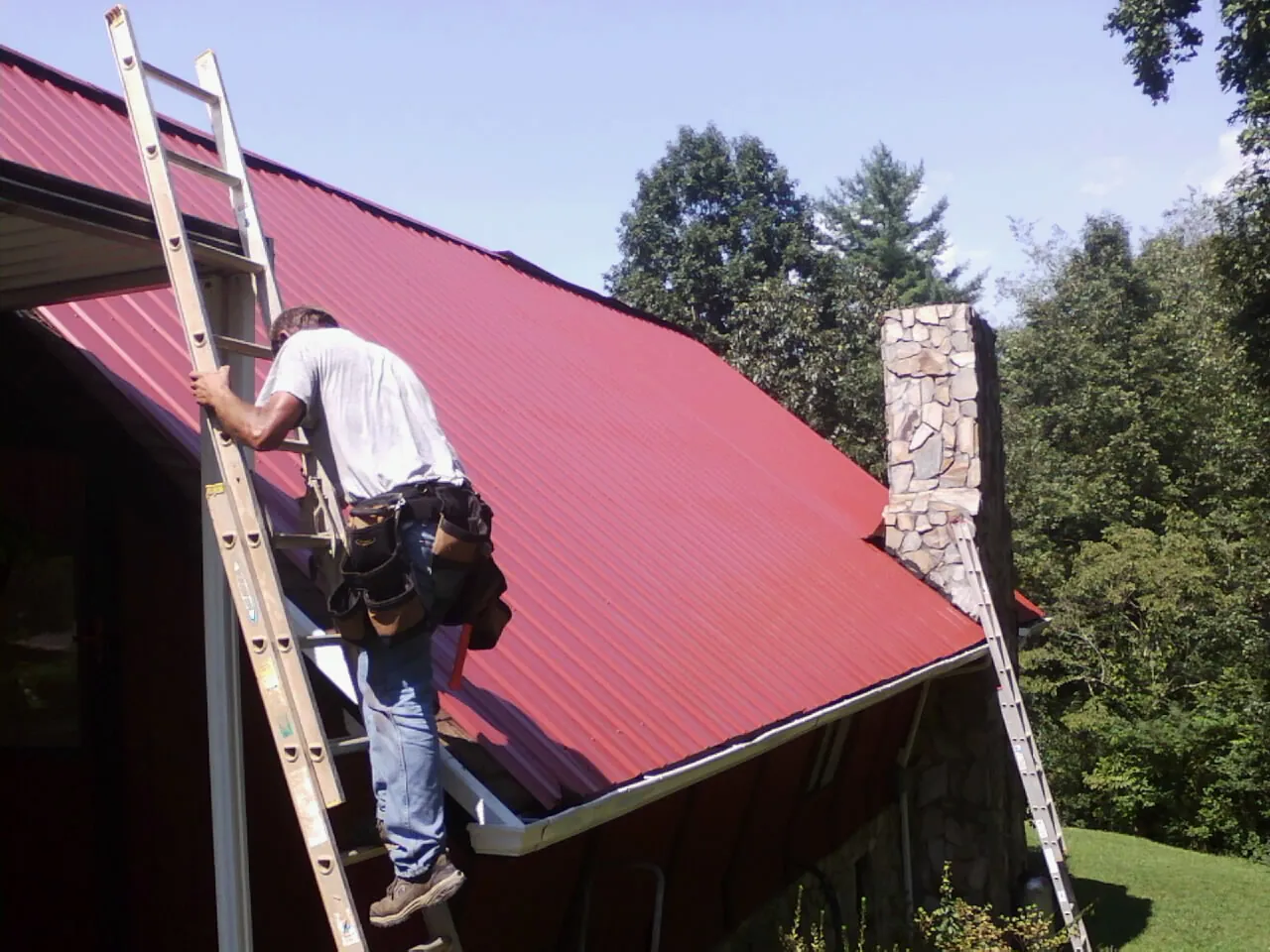 Roofer on a ladder beside a red metal roof and stone chimney — Soffit & Fascia Repair by Roofing Brothers in Eva